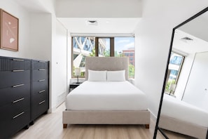 Ein modernes Schlafzimmer mit einem Bett, einer Kommode und einem großen Fenster mit Blick auf Gebäude.
