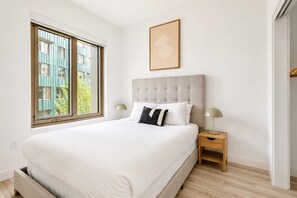 Ein Schlafzimmer mit einem großen Bett, einem Fenster mit Blick auf Gebäude, einem Nachttisch mit einer Lampe und einem gerahmten Bild an der Wand.
