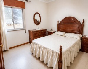 Ein Schlafzimmer mit einem großen Bett, einem Holz-Kopfteil, einem Schminktisch, einem Nachttisch, einem Spiegel und einem Fenster mit Jalousien.