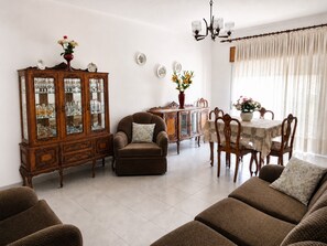 Ein Wohnzimmer mit braunem Sofa, einer Holzvitrine mit Glastüren, einem Esstisch mit Stühlen und einer Kronleuchter.