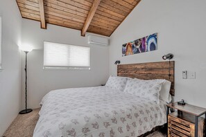 Un dormitorio con techo de madera, una cama con ropa de cama floral, una lámpara de pie, una ventana con persianas y un aire acondicionado de pared.