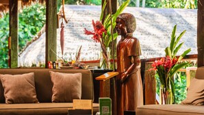 Eine Holzstatue, ein Sofa und ein Tisch mit einer Blumenvase in einem Raum, dessen Dach mit Stroh gedeckt ist und durch viel Grün gekennzeichnet ist.
