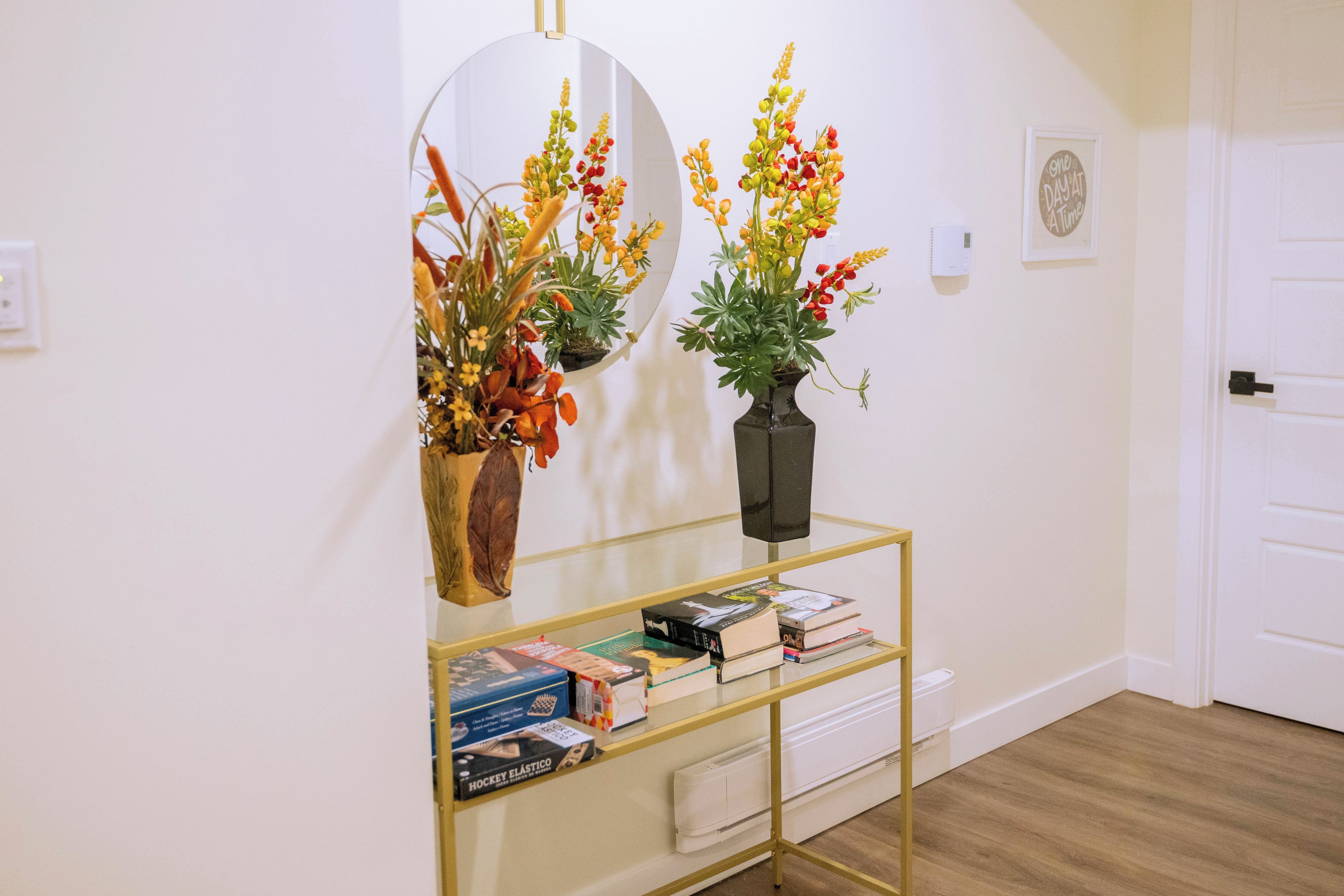 A hallway with a round mirror, a vase with flowers, a glass shelf with books, and a door.