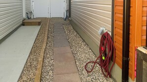 Een smal pad, begrensd door gebouwen met beplating en een houten deur, dat leidt naar een hekwerk.