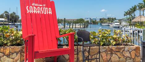 Uma cadeira vermelha com a inscrição 'Welcome to Sarasota', uma parede de pedra e um porto com barcos.