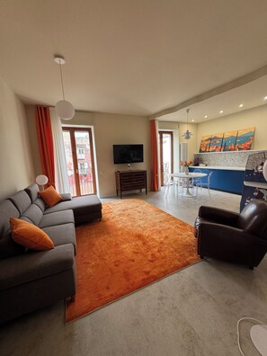 A living room with a sofa, a coffee table, a TV, and a kitchen area with a dining table. - Napolispacca Holiday Home by H4H (Napoli)