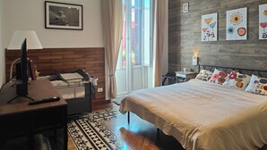 Een slaapkamer met een bed, een bureau, een stoel, een televisie, een raam met gordijnen en kunst aan de muur.