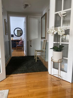 A hallway with a black rug, a wooden chair, a gold-framed chair, a white potted plant, and a mirror on the wall. - Warm little jewels! (Longueuil)