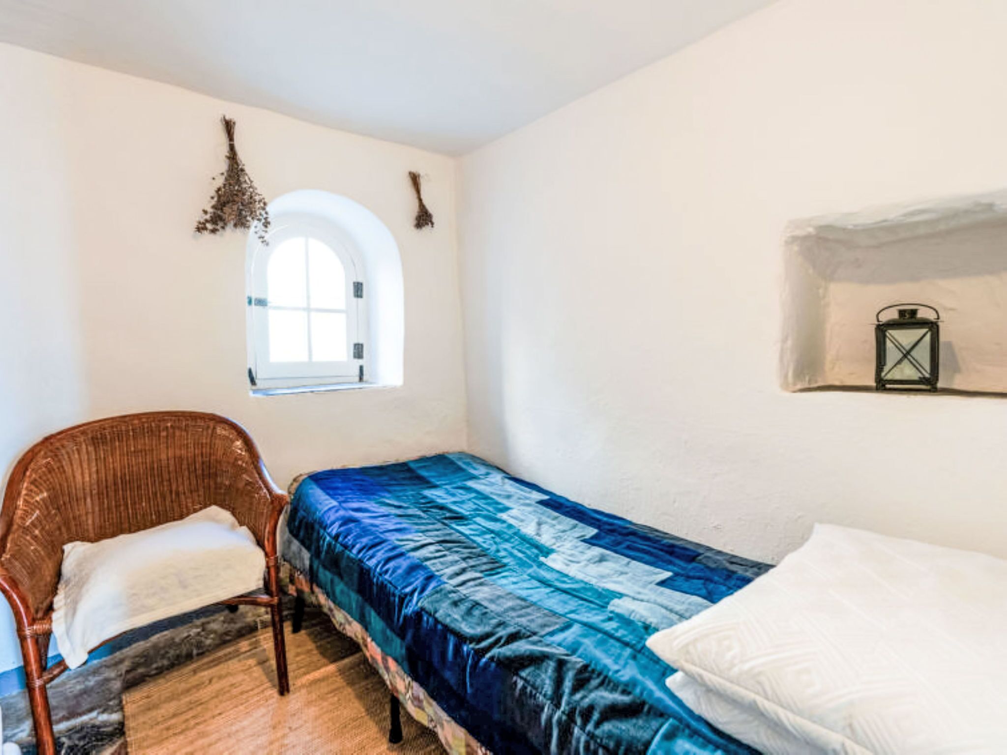 Een kleine kamer met een eenpersoonsbed, een stoel, een raam en een wandplankje met een lantaarn.