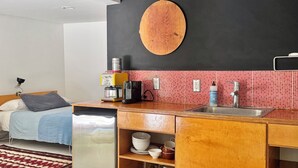 A compact kitchen with a sink, countertop, and storage cabinets. A bed with a blue blanket and pillows is visible in the background. - Fantastic studio with AC in vibrant Los Angeles neighborhood. (Los Angeles)