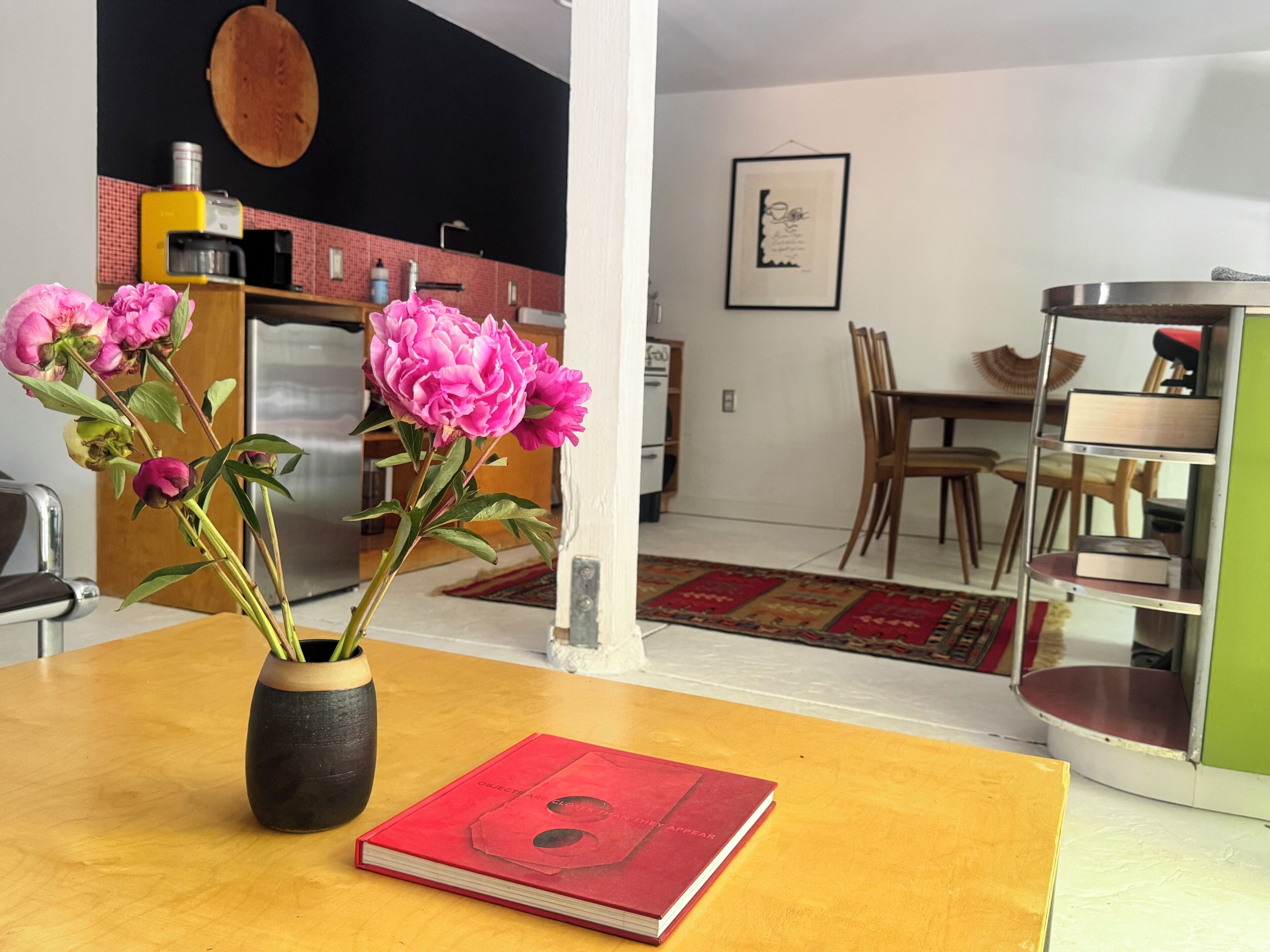 Een kamer met een houten tafel, een vaas met bloemen, een rood boek, een keuken op de achtergrond en een eetgedeelte met stoelen en een tafel.