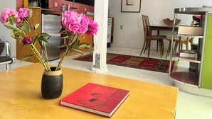 A room with a wooden table, a vase with flowers, a red book, a kitchen in the background, and a dining area with chairs and a table. - Fantastic studio with AC in vibrant Los Angeles neighborhood. (Los Angeles)