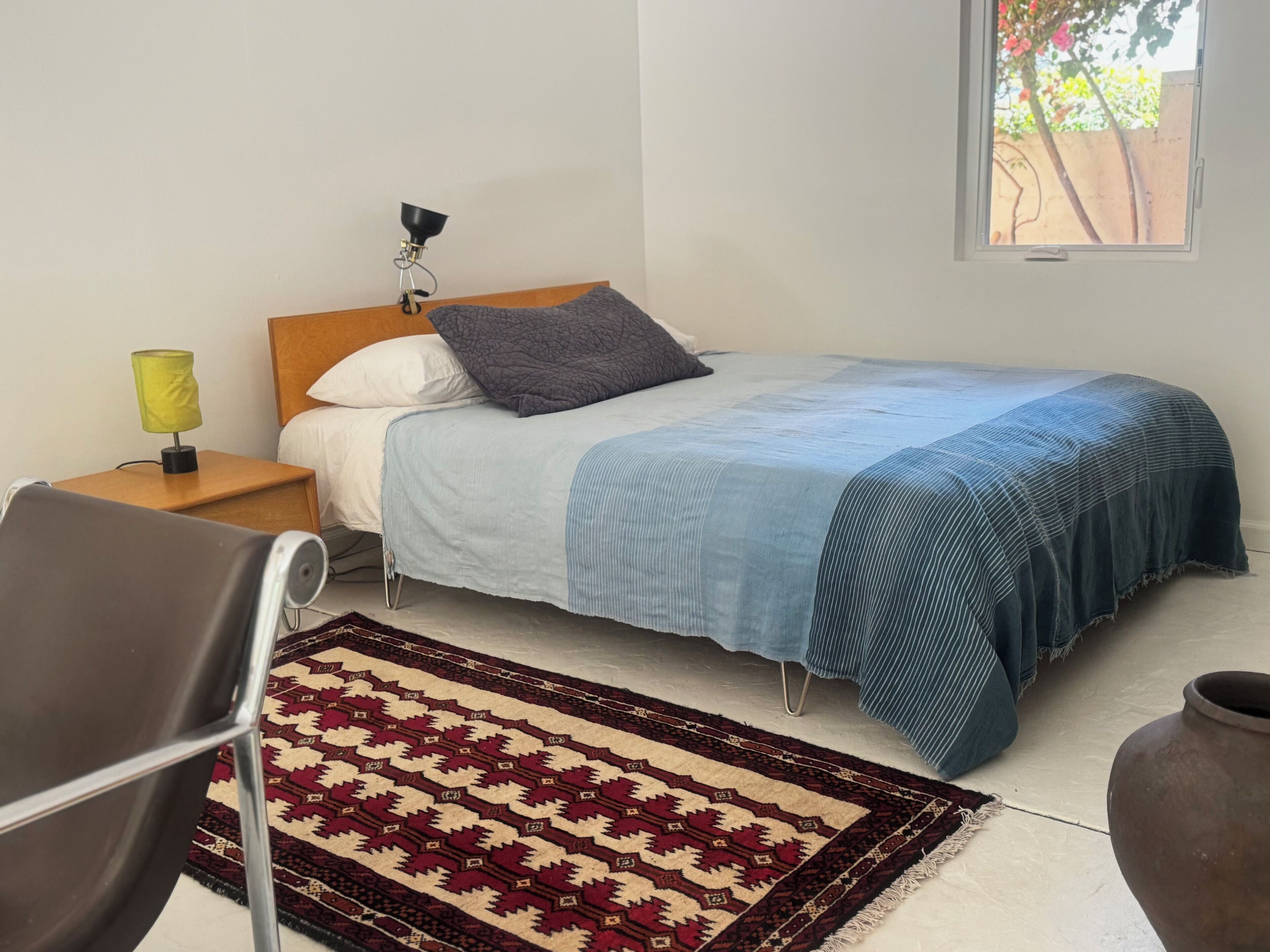 Ein Schlafzimmer mit Bett, Nachttisch mit Lampe, Stuhl, Teppich mit Muster und Fenster.