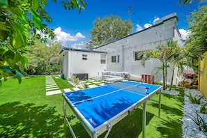 Ein Pingpongtisch in einem Garten mit einem weißen Haus, einer grünen Rasenfläche und einem Schachbrettweg.