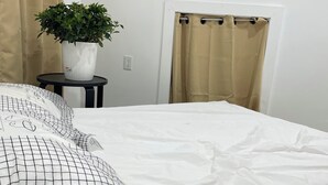 A neatly made bed with a white sheet and a black grid-patterned pillow, a potted plant on a small table, a television mounted on the wall, and a shower curtain. - Private room in tiny home in Brooklyn, New York (Brooklyn)