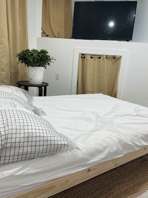 A neatly made bed with a white sheet and a black grid-patterned pillow, a potted plant on a small table, a television mounted on the wall, and a shower curtain. - Private room in tiny home in Brooklyn, New York (Brooklyn)