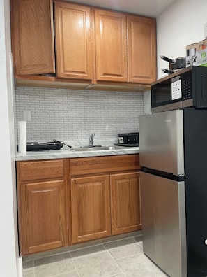 A kitchen with wooden cabinets, a stainless steel refrigerator, a microwave, and a tiled backsplash. - Private room in tiny home in Brooklyn, New York (Brooklyn)