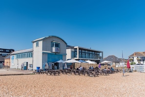 Un edificio sul lungomare con posti a sedere all'aperto e ombrelloni, cielo sereno e spiaggia di ciottoli.