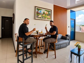 Two individuals seated at a table with plates of food, a painting on the wall, and a view of the outside through a door.