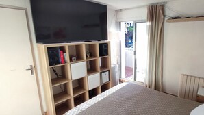 Quarto de hotel com cama, televisão, escrivaninha com luminária, radiador e janela com cortinas.