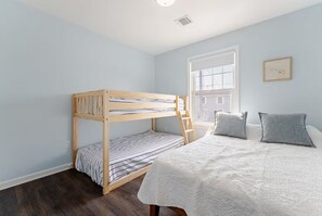 Een kamer met een stapelbed, een raam met jaloezieën, een nachtkastje en een bed met kussens.