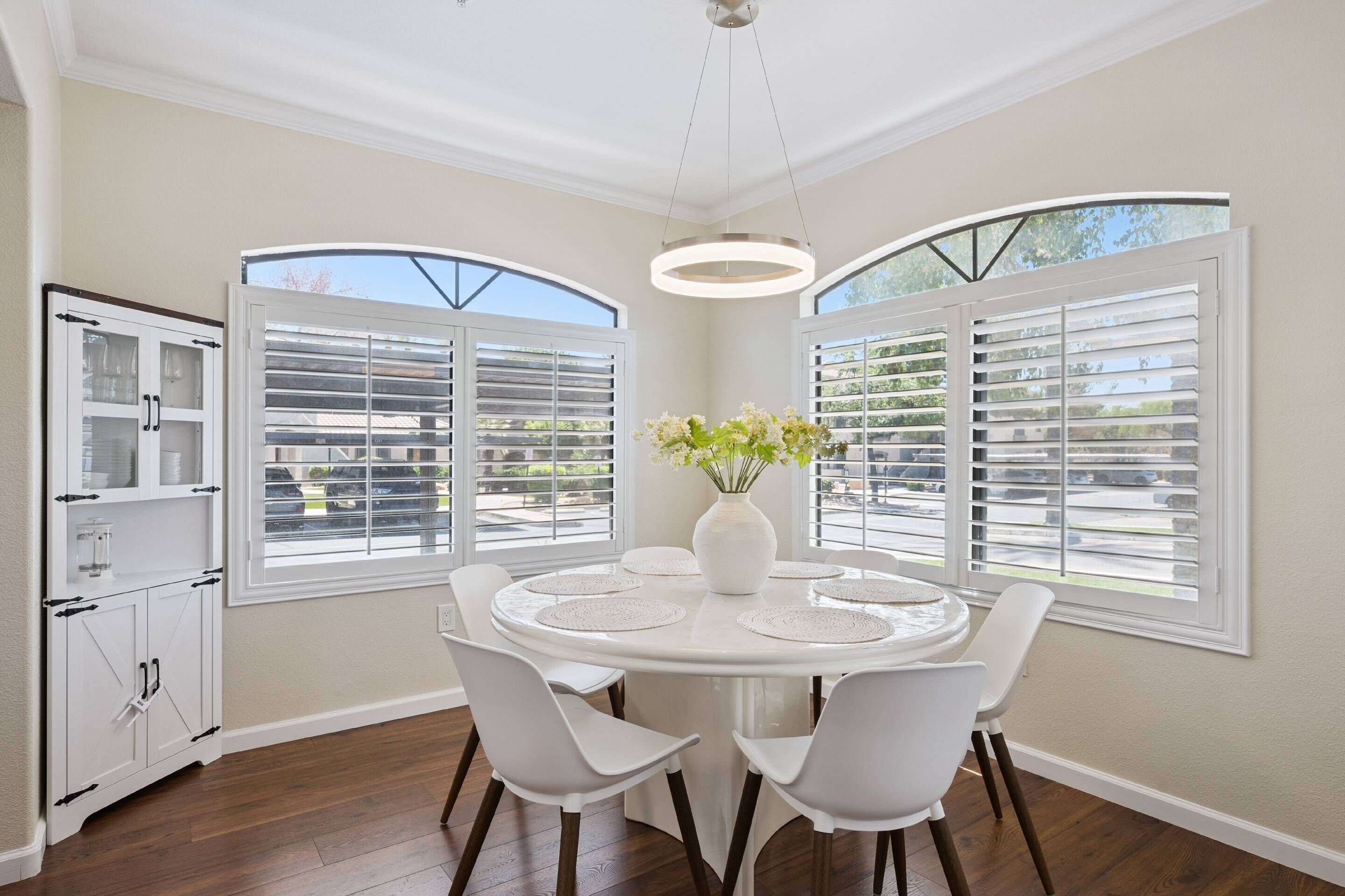 A dining area with a round table, white chairs, a vase with flowers, and large windows with shutters.