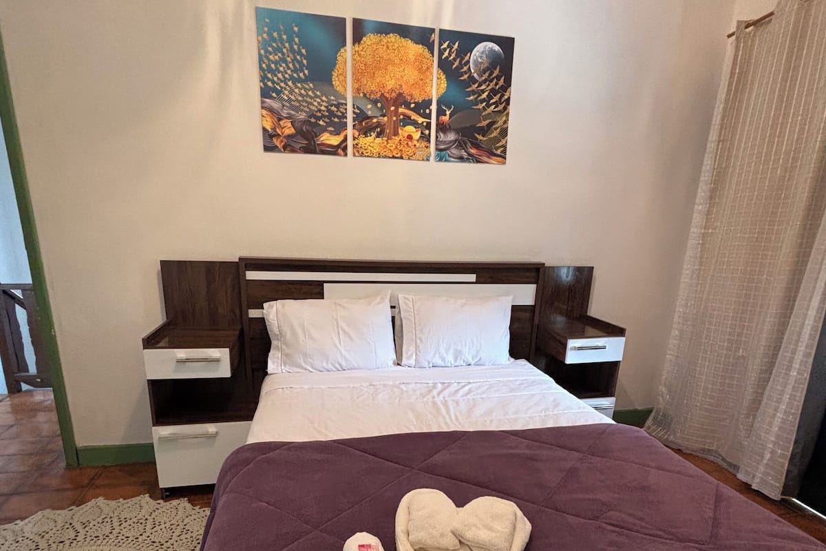 A hotel room with a bed, two bedside tables, a nightstand, a large painting, and a window with curtains.