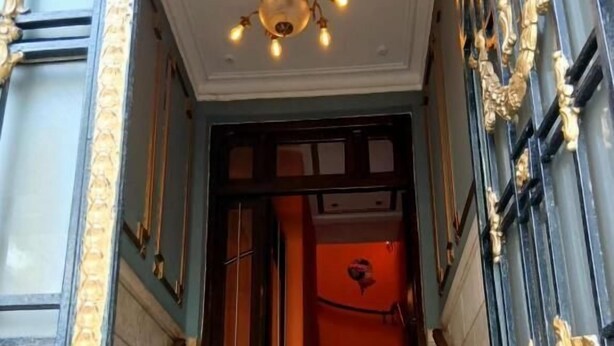 Una imponente escalera con barandales ornamentados en negro y dorado, un candelabro y una pared roja en la parte superior.