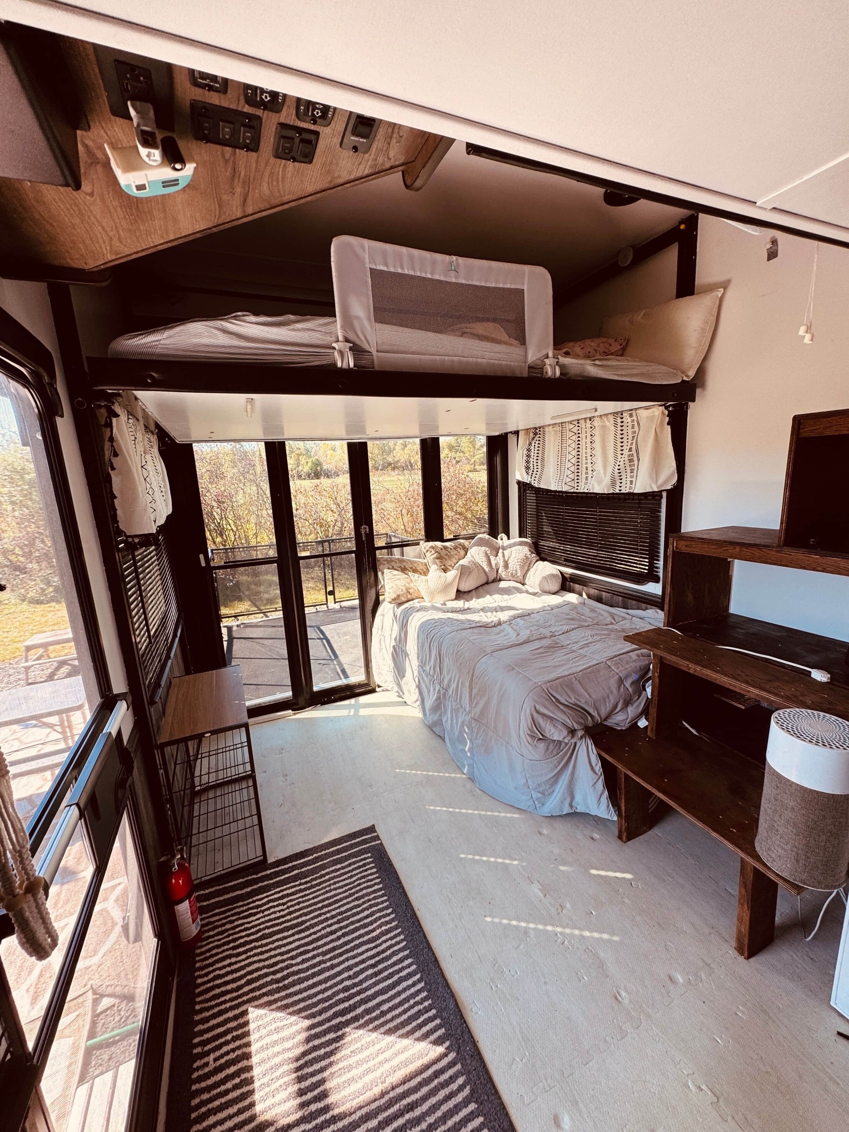 A compact RV interior with a bunk bed, a small desk, and a window providing a view of the outdoors.