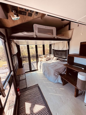 A compact RV interior with a bunk bed, a small desk, and a window providing a view of the outdoors.