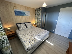 A bedroom with a wooden wall, a bed with patterned bedding, a ceiling light, and a door. - LES EMBRUNS - VIELLE SAINT GIRONS (VIELLE SAINT GIRONS)