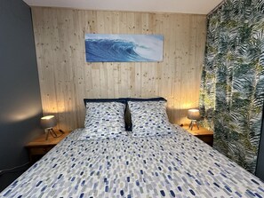 A bedroom with a wooden wall, a bed with patterned bedding, two bedside lamps, and a wall-mounted painting of a wave. - LES EMBRUNS - VIELLE SAINT GIRONS (VIELLE SAINT GIRONS)