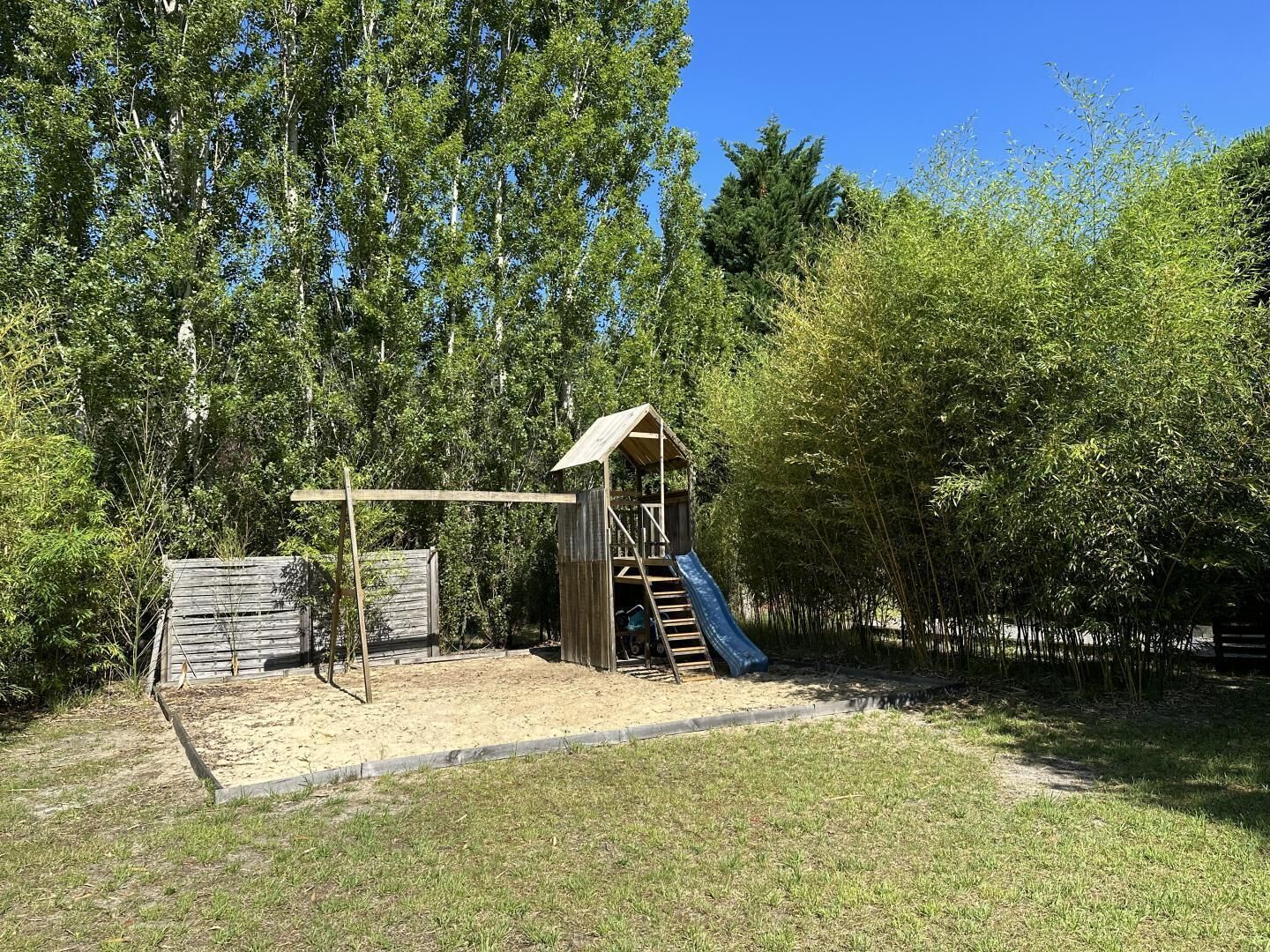 A playground with a slide and climbing structure, surrounded by trees and a sandy area.