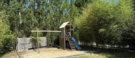 A playground with a slide and climbing structure, surrounded by trees and a sandy area.