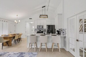 A modern kitchen with white cabinetry, a central island with bar stools, and a dining area with a wooden table and chairs. - Cozy Colony Cottage (The Colony)