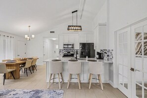 A modern kitchen with white cabinetry, a central island with bar stools, a dining area with a wooden table and chairs, and a patterned rug on the floor. - Beautiful updated home in the heart of The Colony (The Colony)