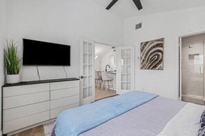 A bedroom with a bed, a TV mounted on the wall, a desk, and a shower area. - Cozy Colony Cottage (The Colony)