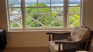Een kamer met een groot raam dat uitzicht geeft op bergen en een meer. Er staat een rieten stoel met een bloemenkussen en een klein wit tafeltje.