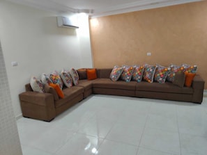 A living room with a brown sectional sofa, colorful patterned pillows, a white tiled floor, and a wall-mounted air conditioning unit.