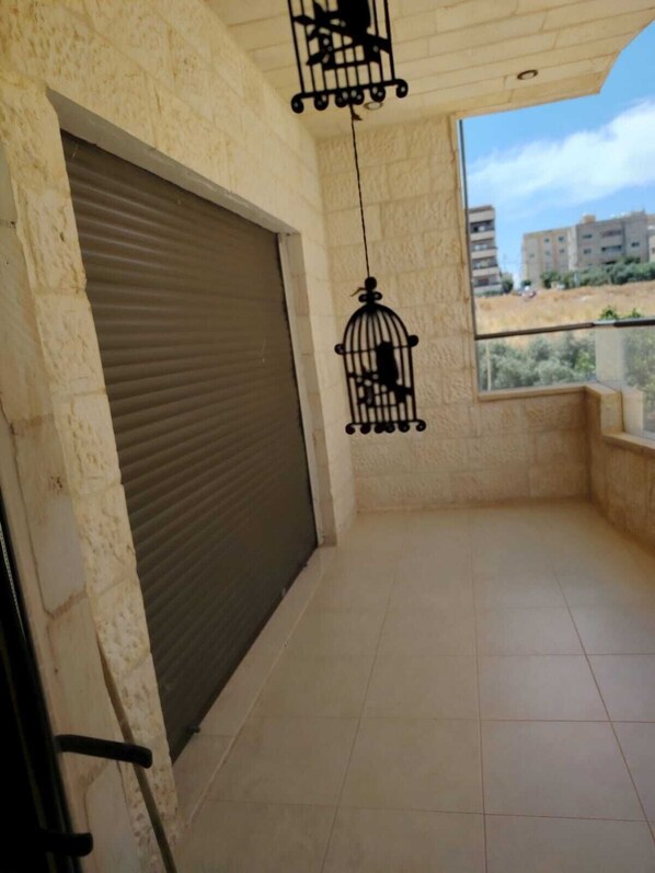 A balcony with a closed shutter, a hanging birdcage, and a view of buildings in the distance.