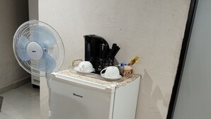 Uma geladeira compacta com uma cafeteira e xícaras em cima, um ventilador e um balde no chão.
