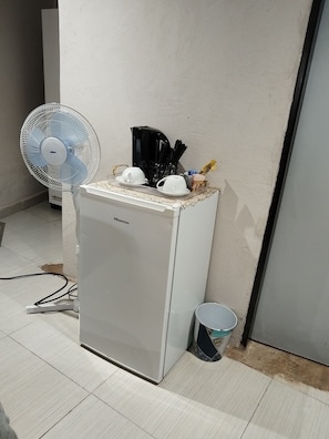 Un petit réfrigérateur, avec une machine à café et des tasses posées sur le dessus, un ventilateur et un seau sur le sol.
