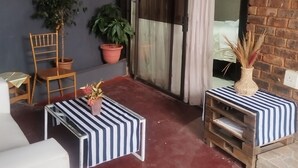 Um pequeno espaço de pátio fechado, com chão vermelho, uma cadeira de madeira, uma mesa pequena com toalhas de mesa listradas, plantas em vasos e uma parede de tijolos.