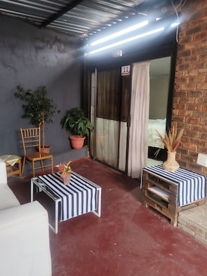 Un petit patio clos, doté d’un sol rouge, d’une chaise en bois, d’une petite table recouverte d’une nappe à rayures, de plantes en pot et d’un mur en briques.