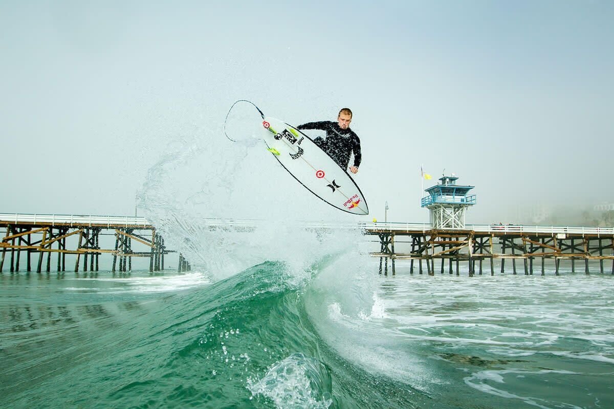 A surfer performing a trick on a wave with a pier in the background.