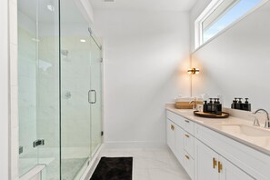 Un baño moderno con cabina de ducha de vidrio, un espejo grande y tocador con lavamanos y artículos de aseo.