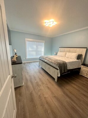 A bedroom with a bed, a dresser, a nightstand, a window with blinds, and a ceiling light fixture. - Stay Salty Cottage (Foley)