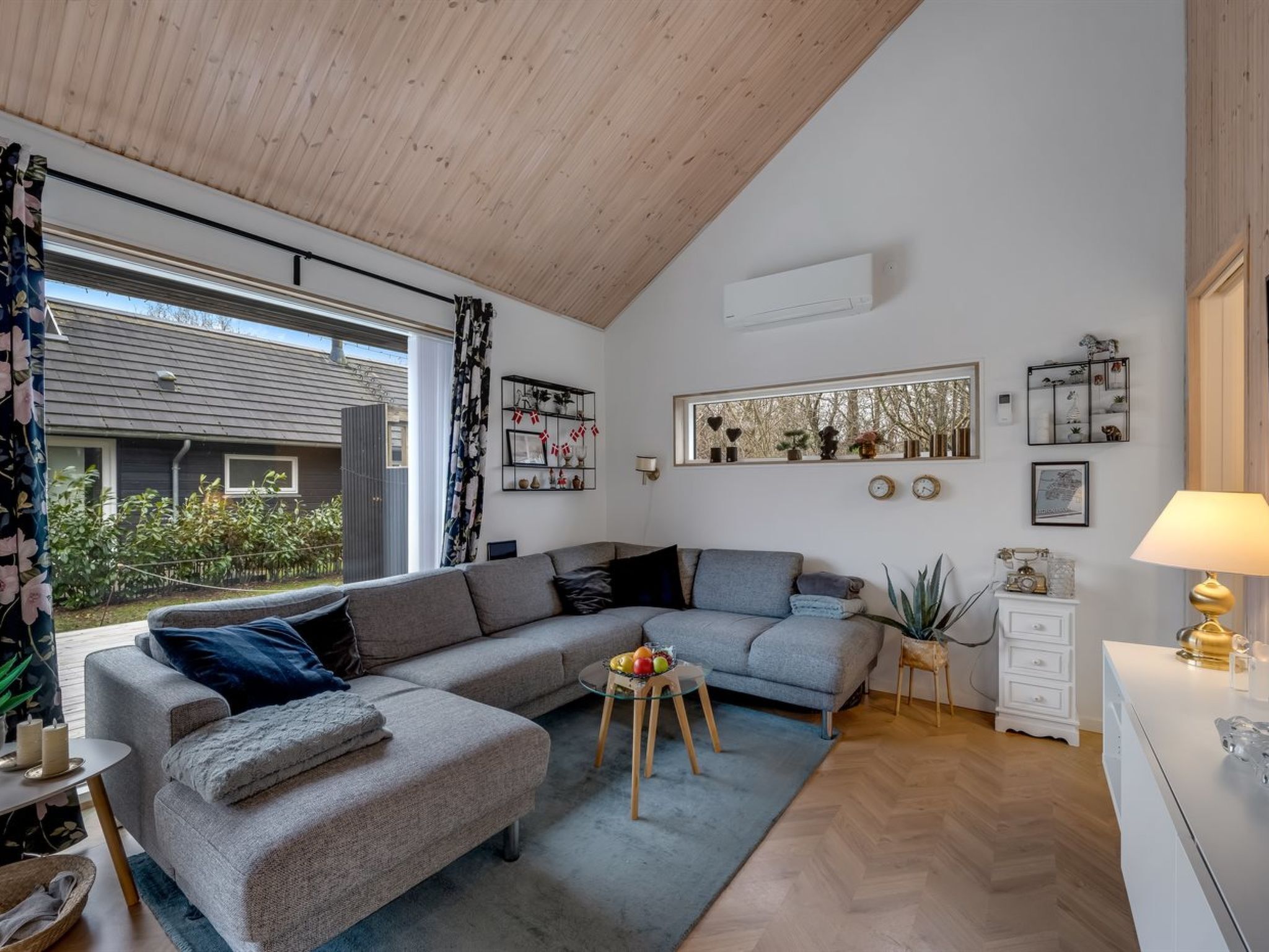 A modern living room with a grey sectional sofa, a small round table with fruit, a white dresser, and a window with a view of greenery.