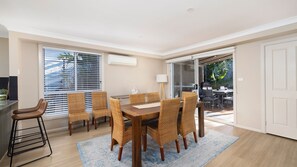 A dining area with a wooden table and wicker chairs, a bar stool, and a sliding glass door leading to an outdoor patio. - Chelsea Poolside and Tennis (Nelson Bay)