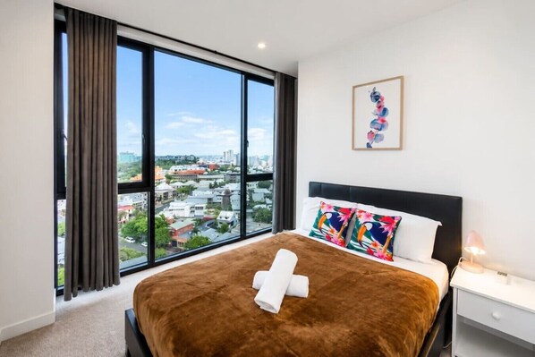 Ein Hotelzimmer mit einem großen Bett, einem Nachttisch, einer Lampe, einem Fenster mit Vorhängen und Blick auf die Stadt.